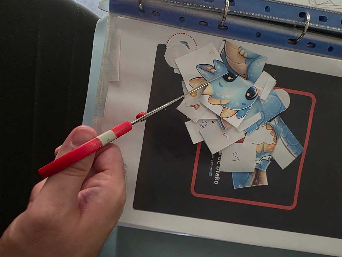 Une main découpe aux ciseaux les pièces du puzzle de Drako, sur la page imprimée d’un livret glissée dans un classeur.