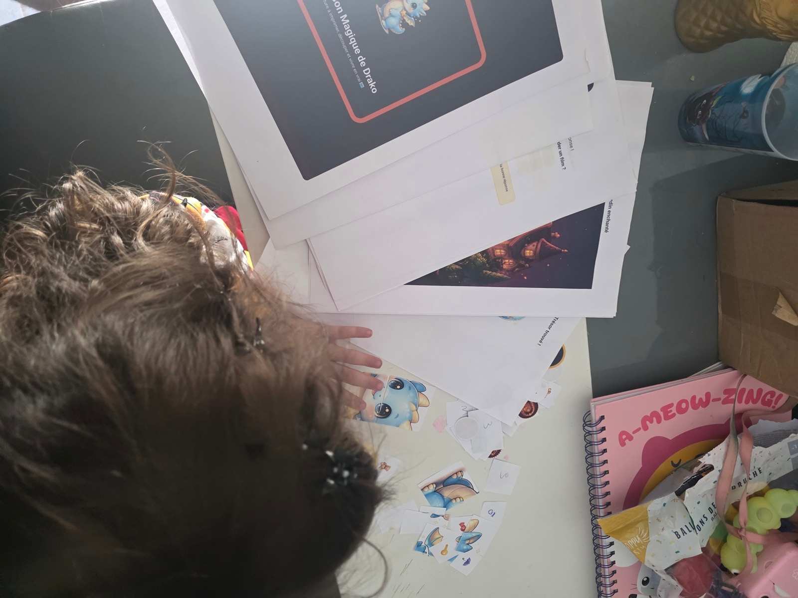 Un enfant penché sur les pages imprimées de « La Maison Magique de Drako », les cartes de jeu déjà découpées étalées devant lui.