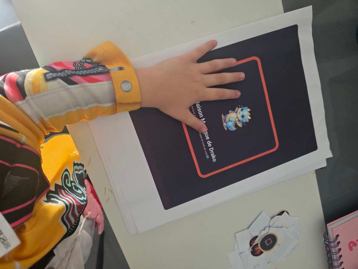La main d’un enfant posée sur la couverture imprimée de « La Maison Magique de Drako », avec les pastilles découpées à côté.